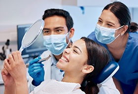 Patient in dentist’s chair smiling at reflection with two dentists looking over her shoulder