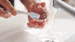 Hands brushing partial dentures at sink with running water